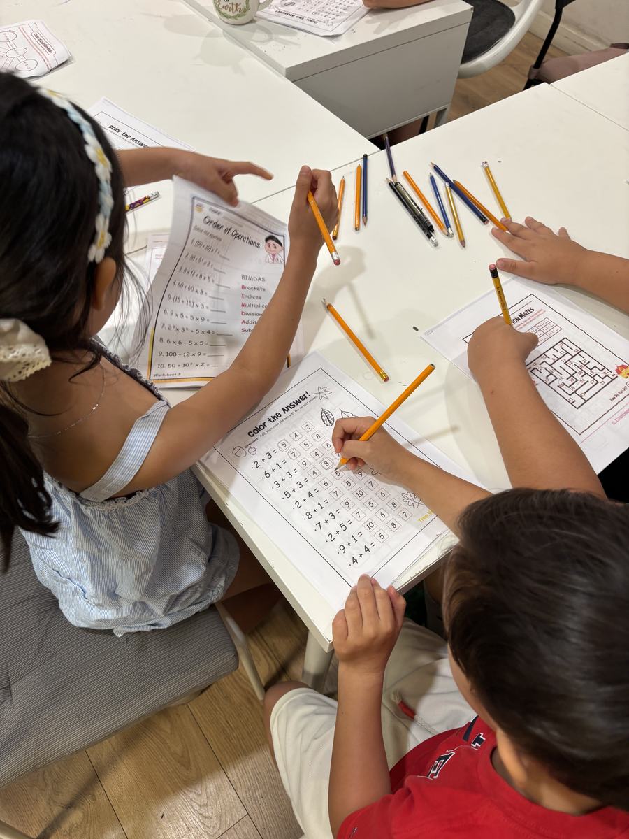 Three different maths worksheets side by side on the table: order of operations, a multiplication maze, and a colour-the-answer addition sheet