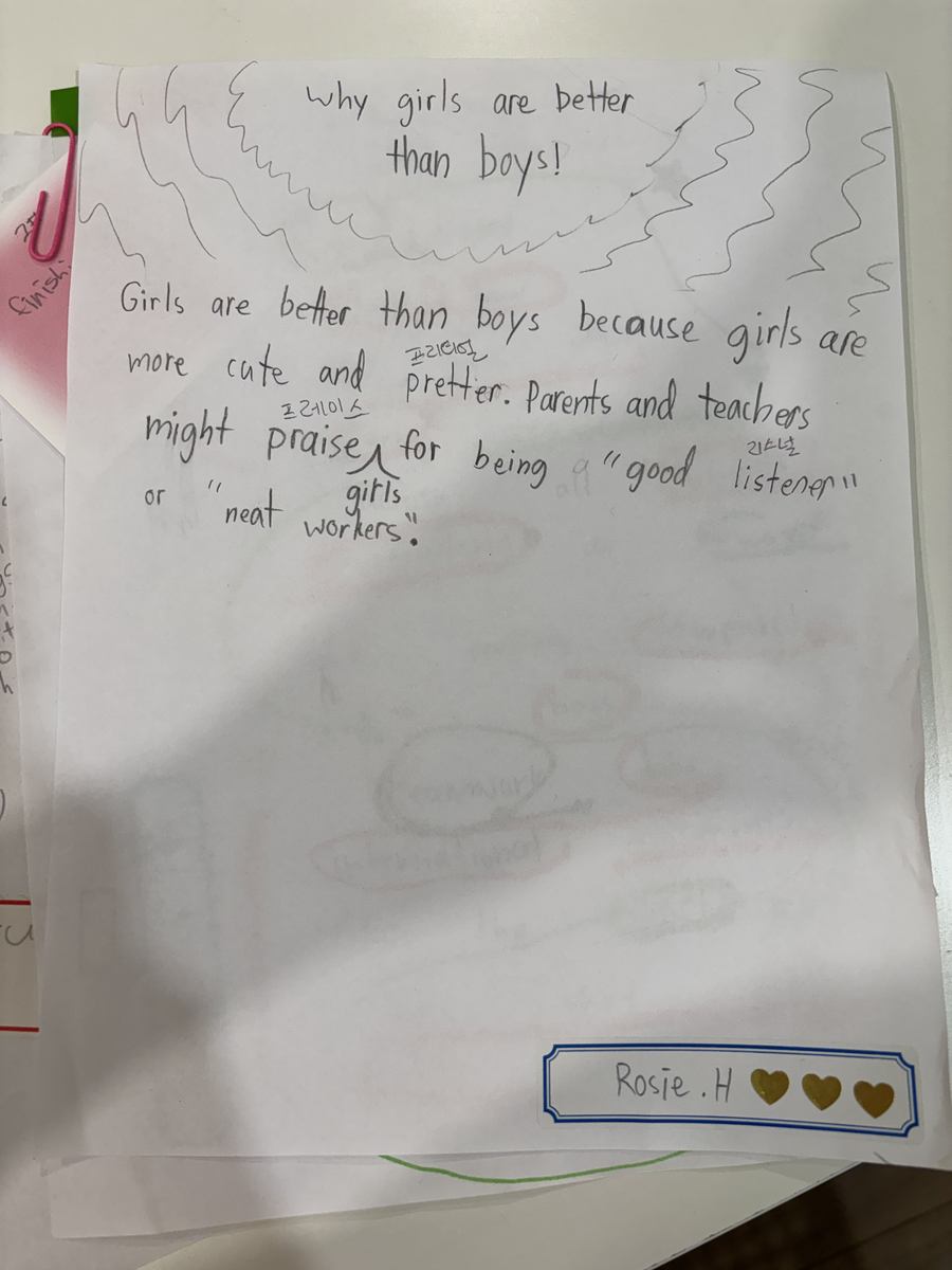 A student's hand-written paragraph with Korean pronunciation notes above the harder English words, signed at the bottom