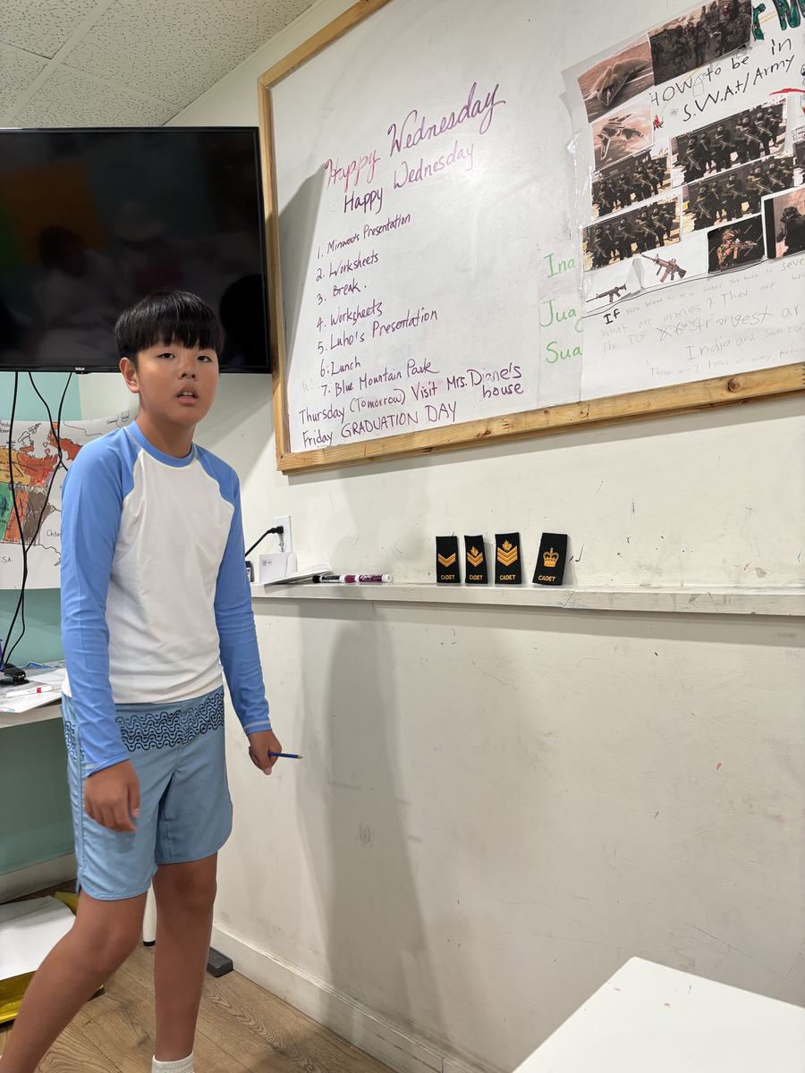Luho standing at the front of the classroom beside his poster on how to be in SWAT and the army, with the day's agenda on the whiteboard