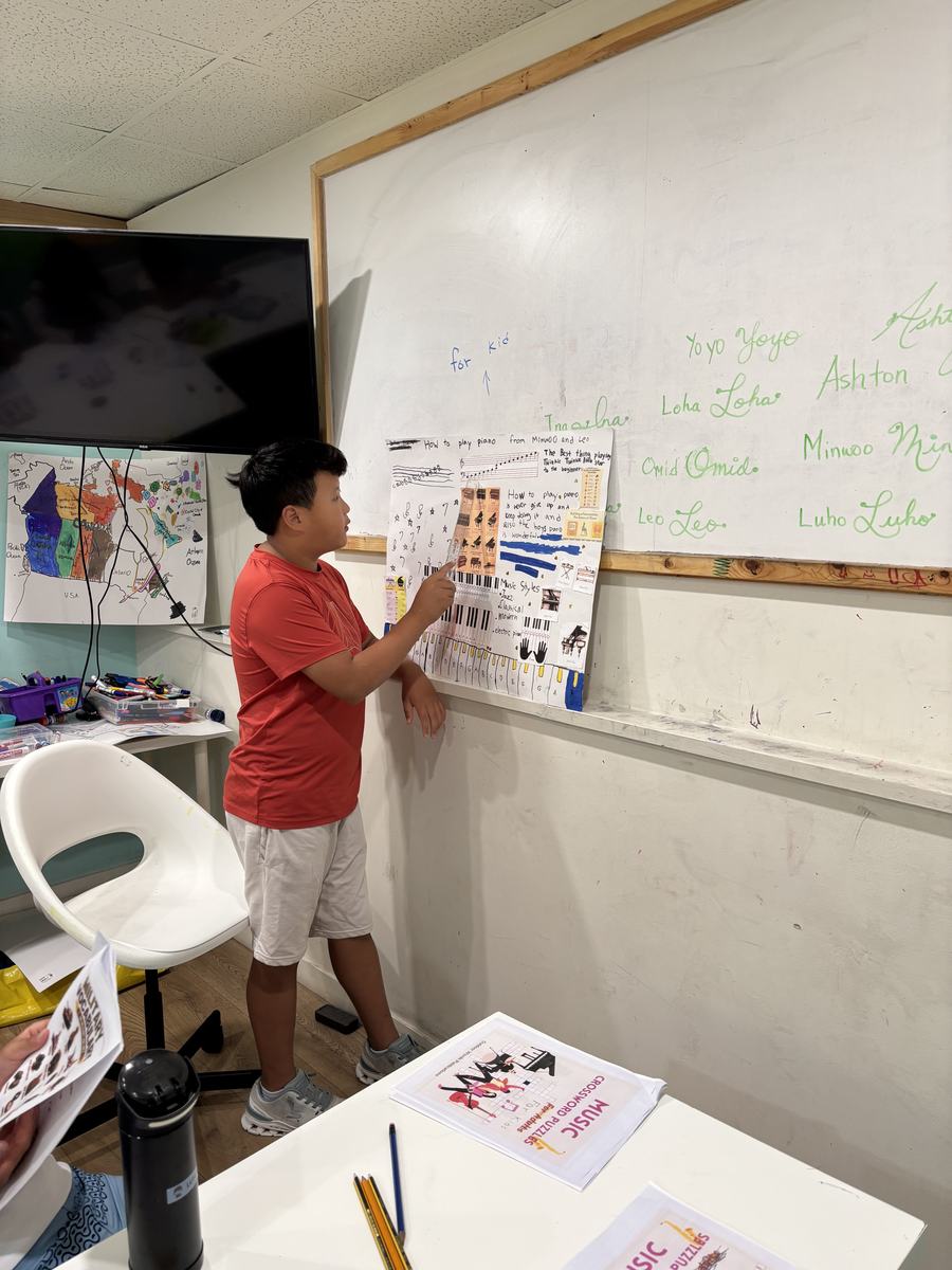 Minwoo standing at the whiteboard pointing to his hand-made piano poster while teaching the class
