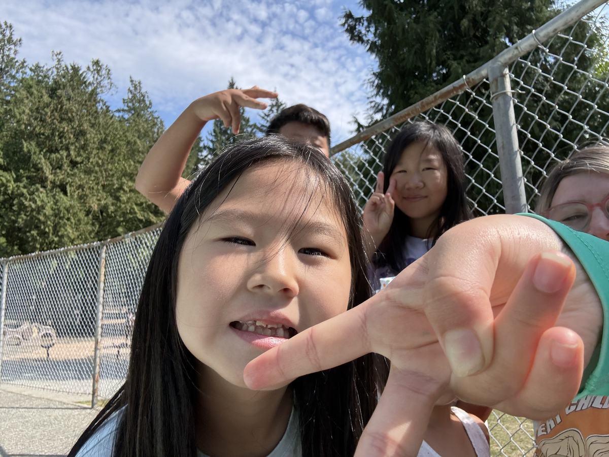 A student leaning into the camera making a peace sign with friends laughing behind her