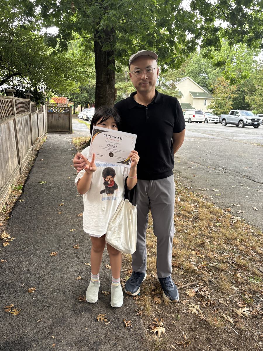 A student holding her certificate up in front of her face beside her father on the driveway