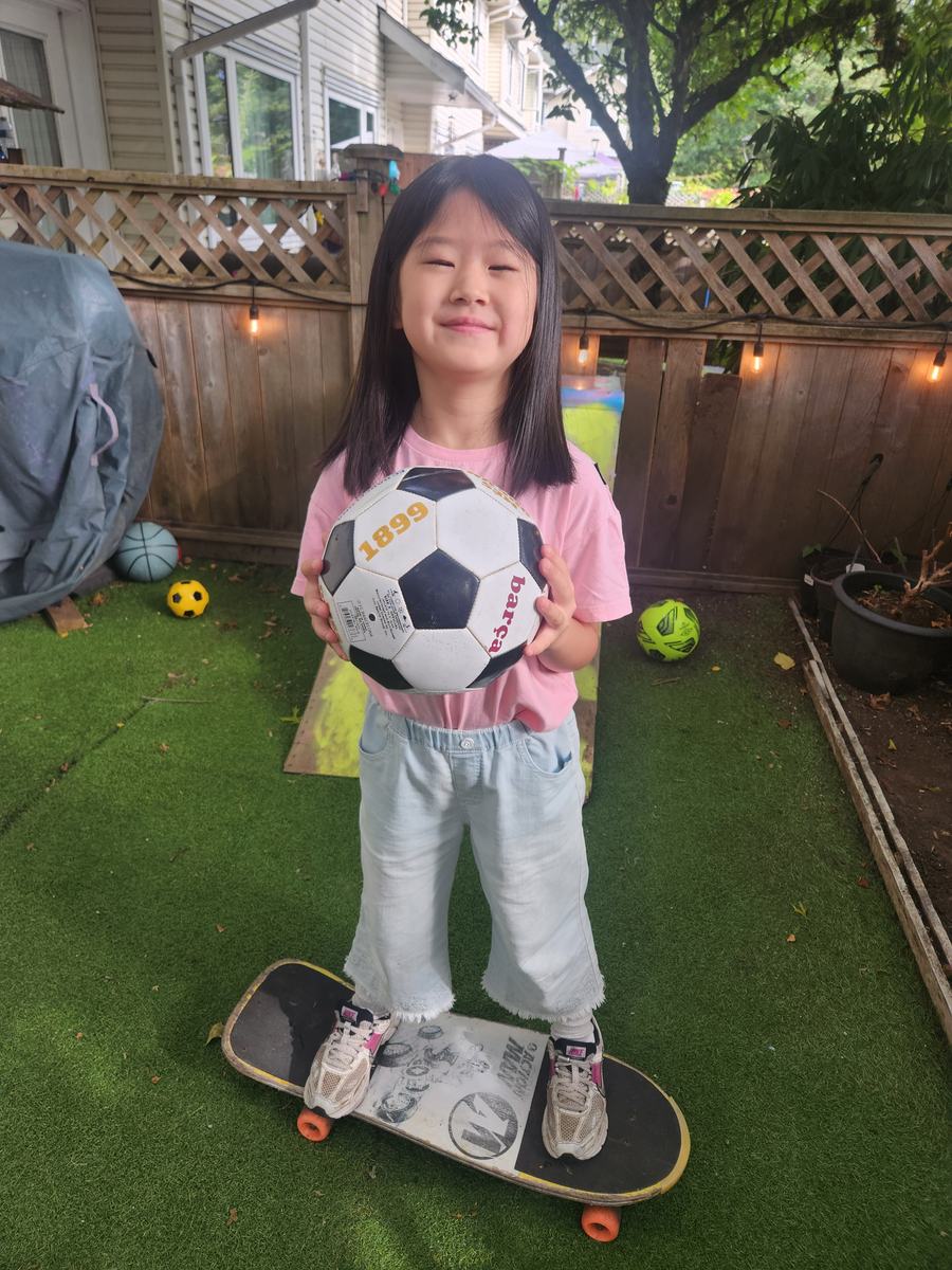 A student standing on a skateboard in the backyard holding a soccer ball