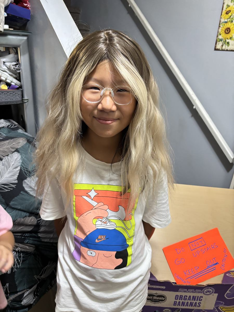 A student wearing the long blonde wig, grinning at the camera indoors
