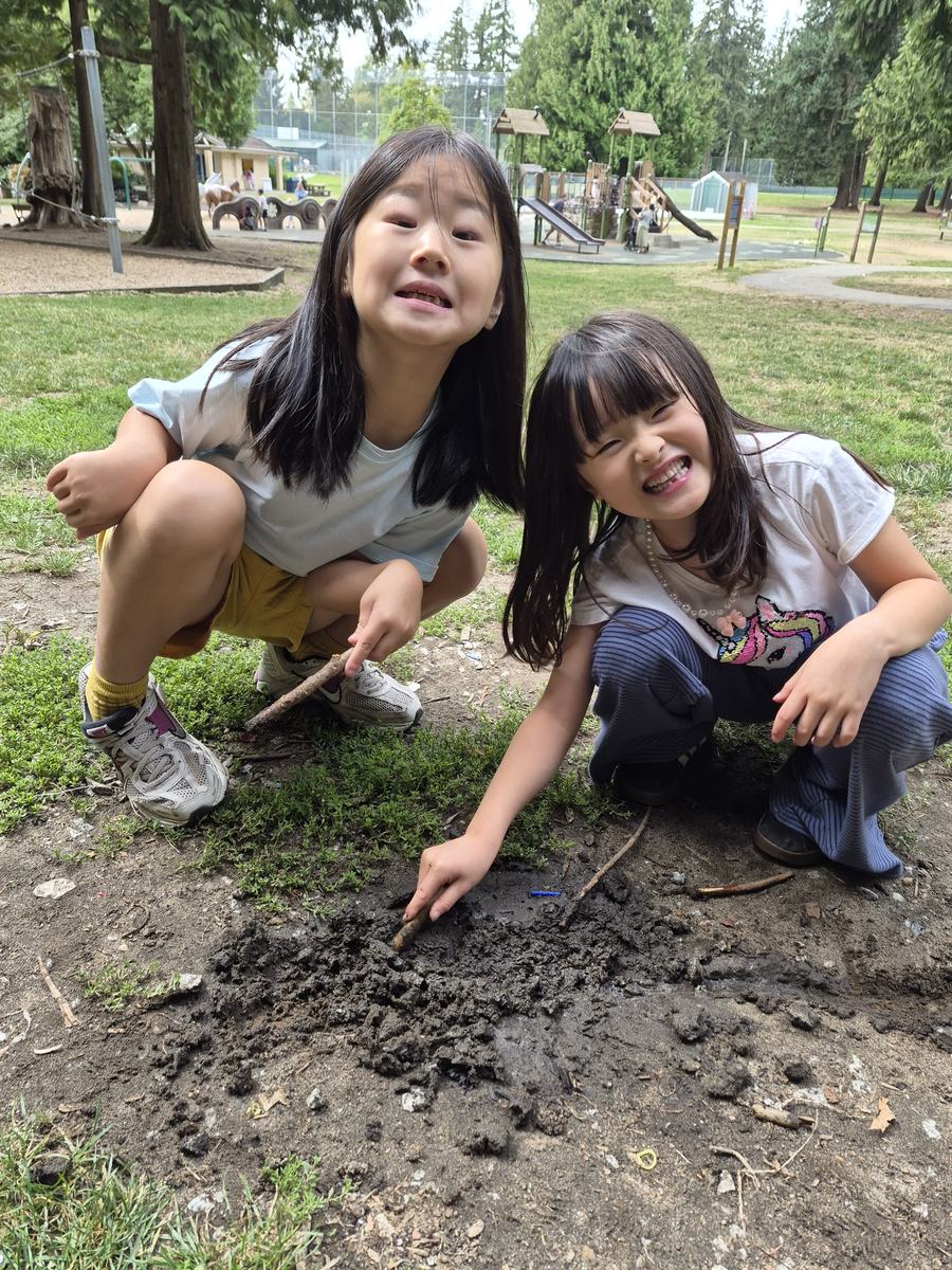 Two students crouched together digging at the dirt with sticks, laughing