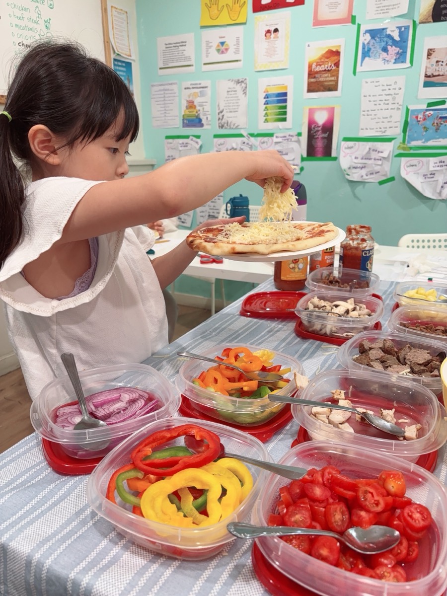 A young student sprinkling grated mozzarella over her finished toppings