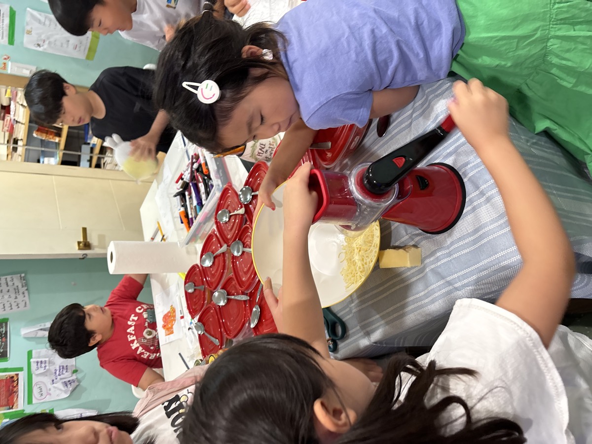 A younger student turning the handle of the cheese grater with help