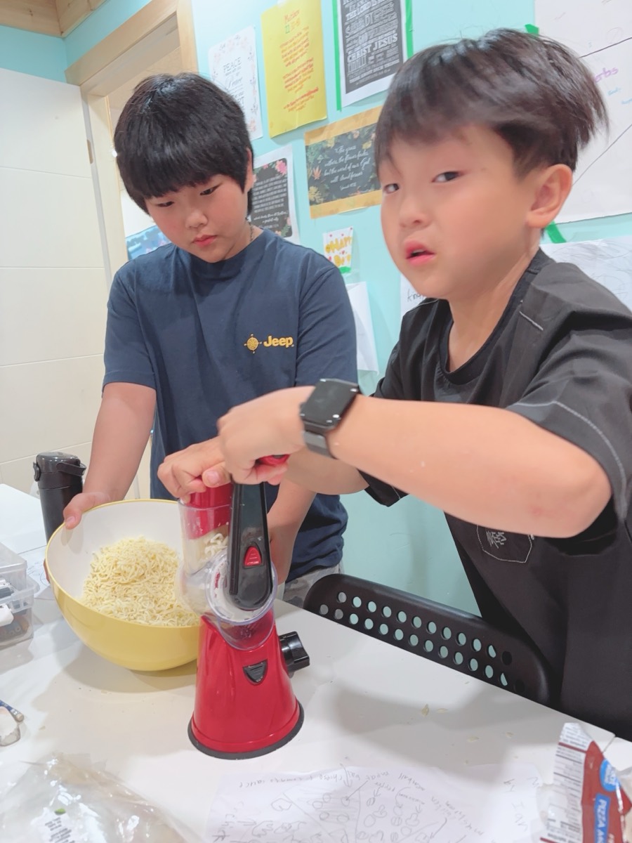 Two students working the rotary cheese grater over a bowl of grated mozzarella