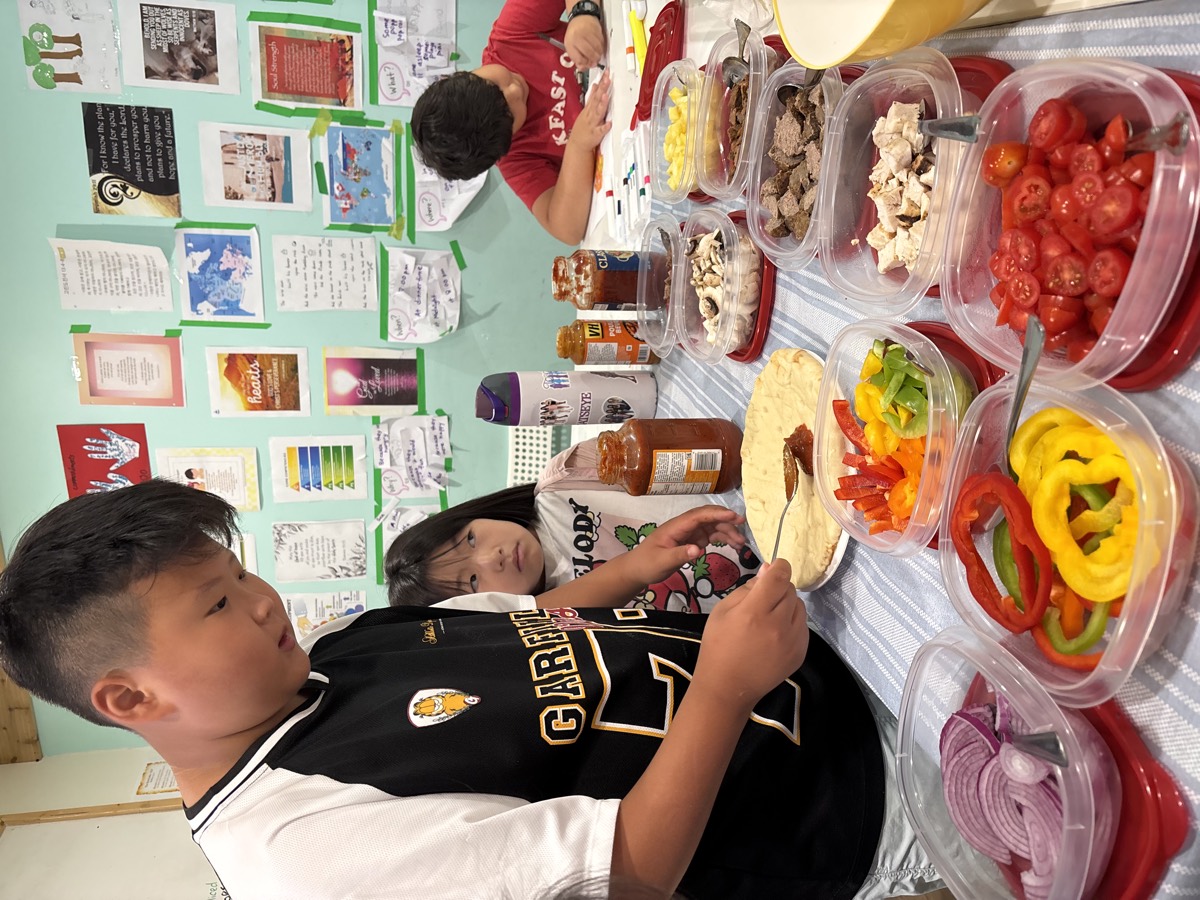 The classroom table covered in containers of colourful pizza toppings while a student assembles his base
