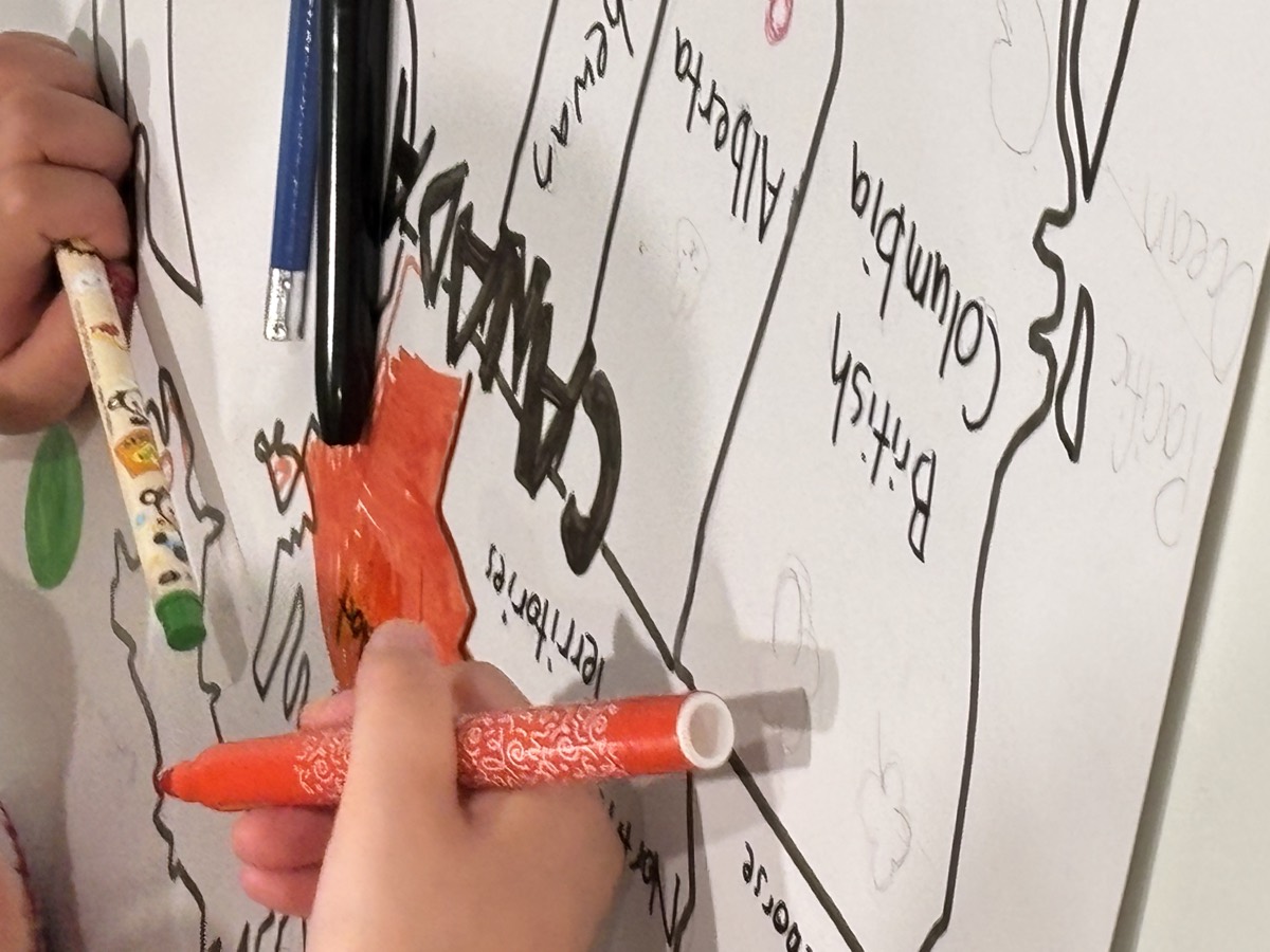 A close-up of a student colouring the word CANADA in red crayon on a large hand-drawn map showing British Columbia and Alberta