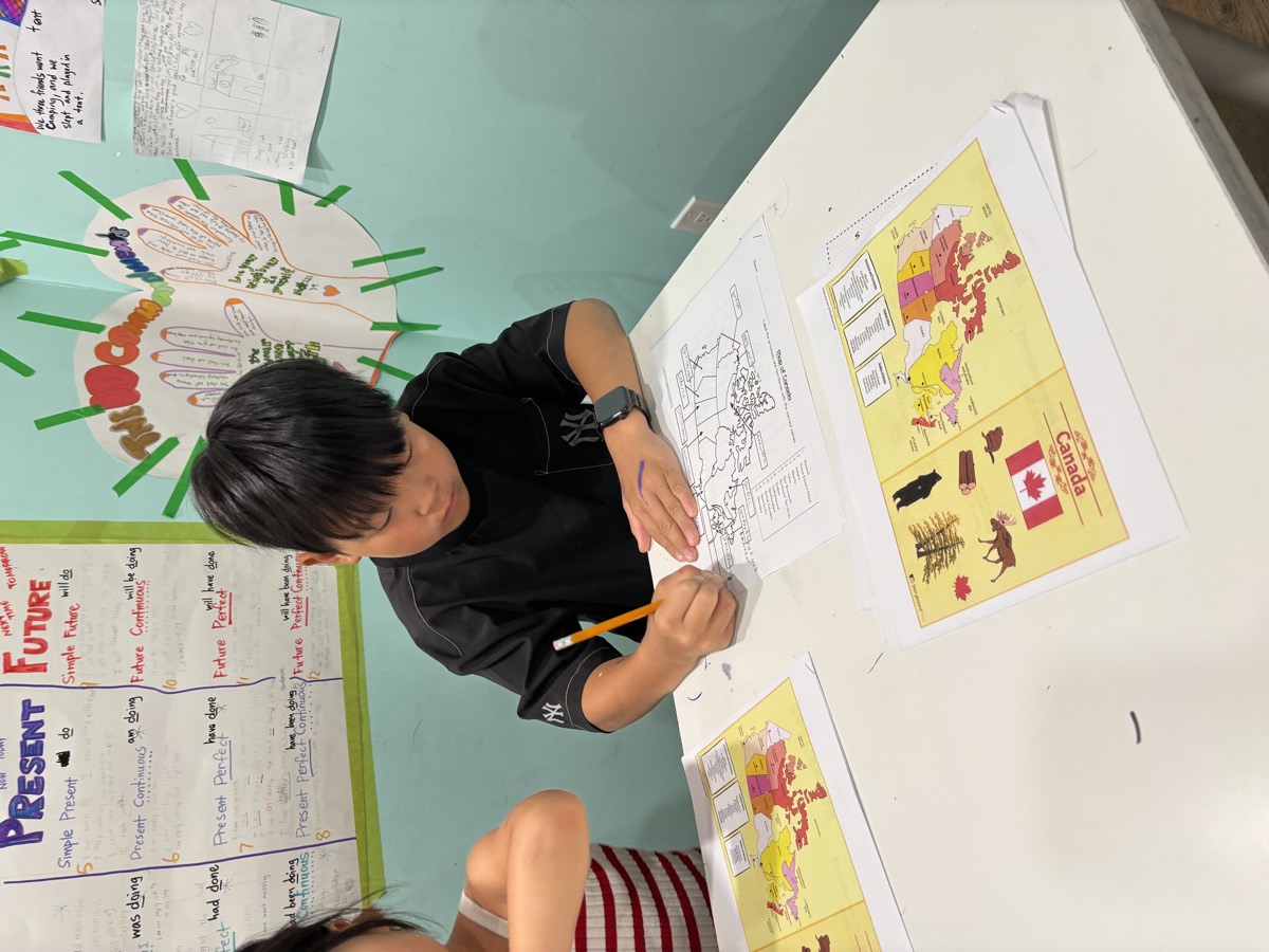 A student labelling a blank map of Canada with a pencil beside a coloured reference map