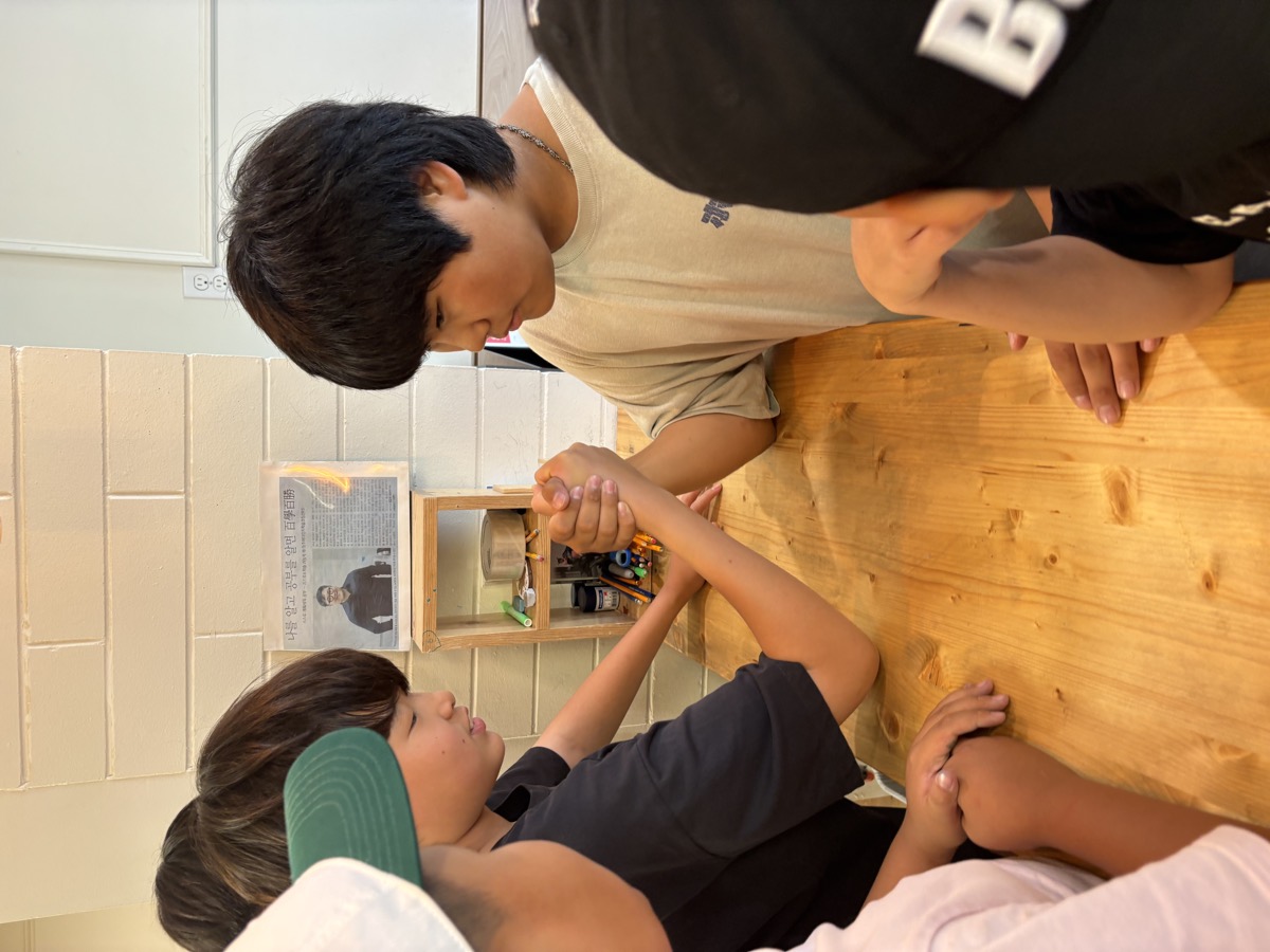 Two older students arm wrestling at the classroom table while friends watch
