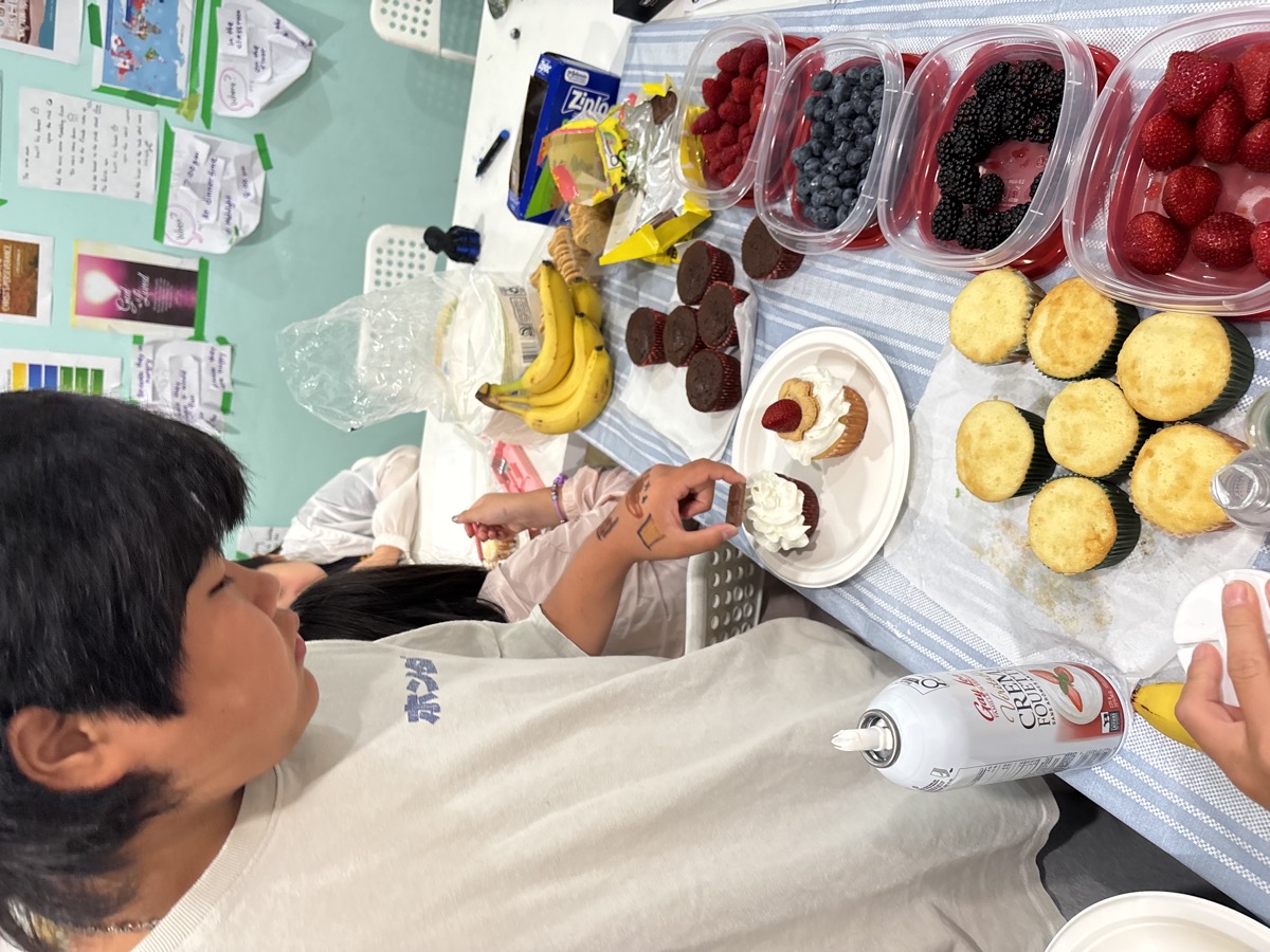 A student carefully adding whipped cream and fruit to his cupcake at the dessert table