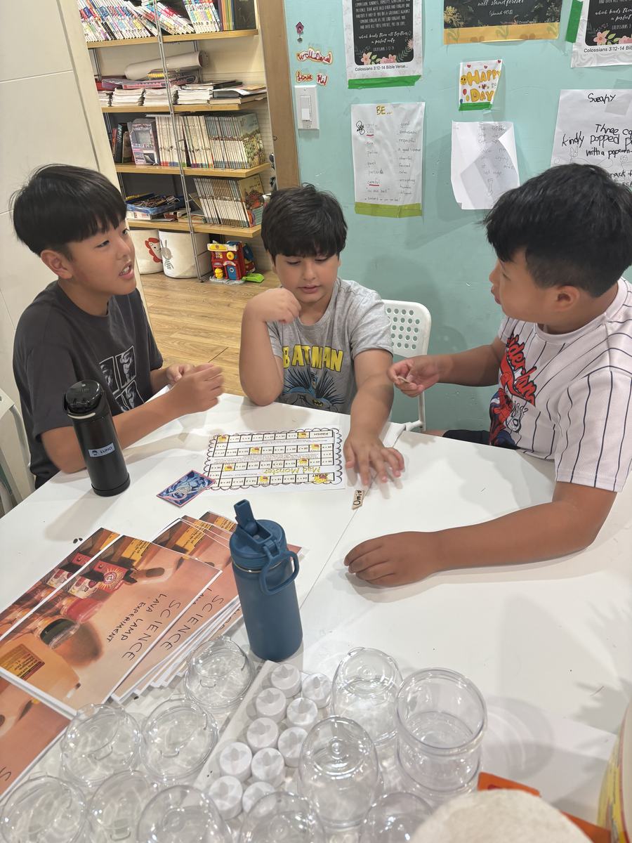 Three boys playing a math board game at the table with the science instruction sheets stacked beside them
