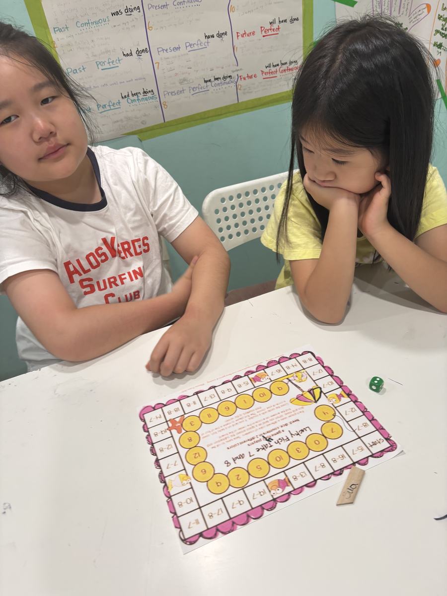 Two students leaning over a printed math board game with a die on the table