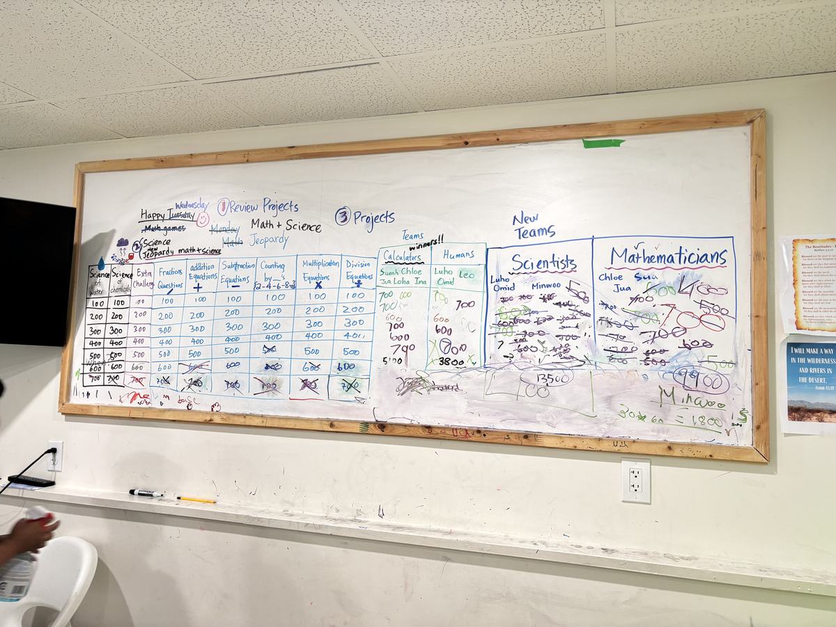 The classroom whiteboard showing the Jeopardy grid extended with science columns, plus both sets of team names and their running scores
