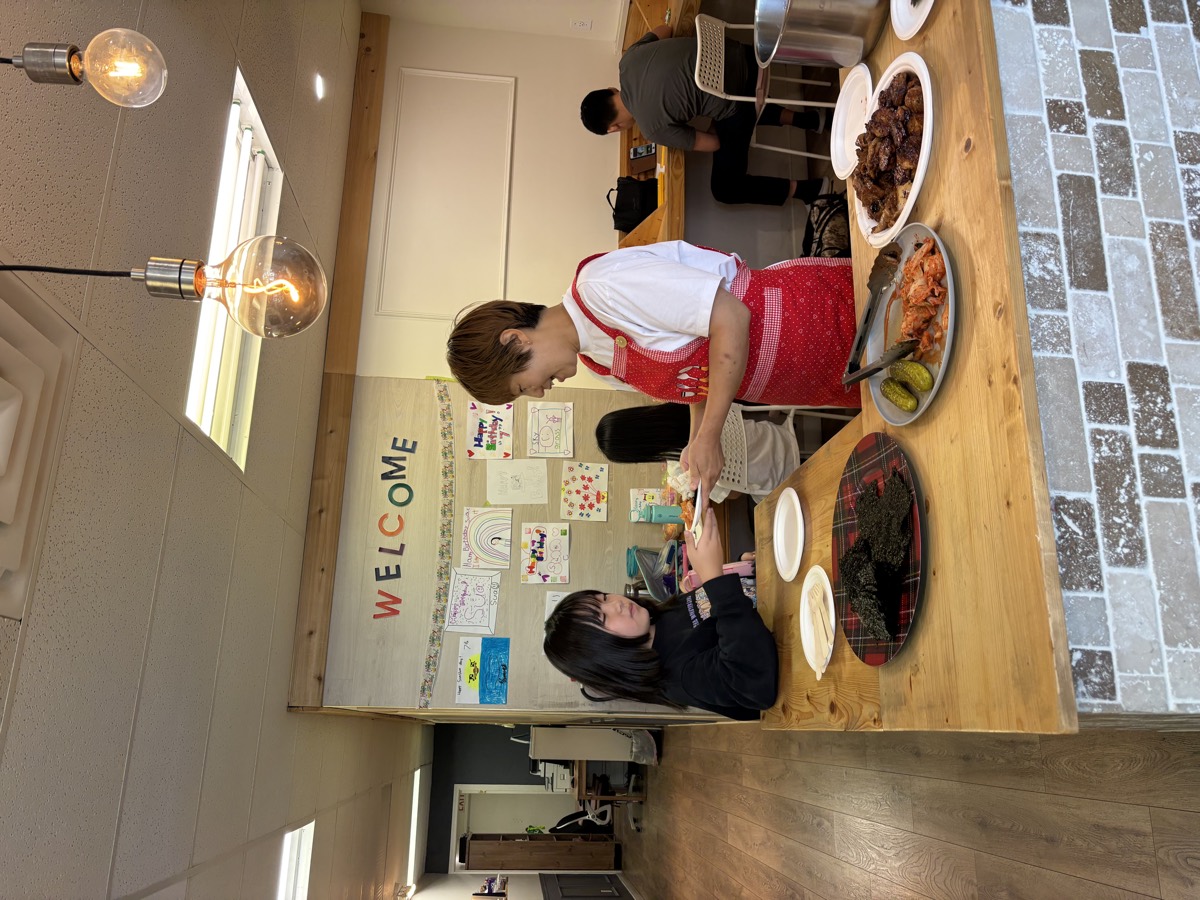 A teacher in a red apron serving a plate of Korean food to a child at the counter