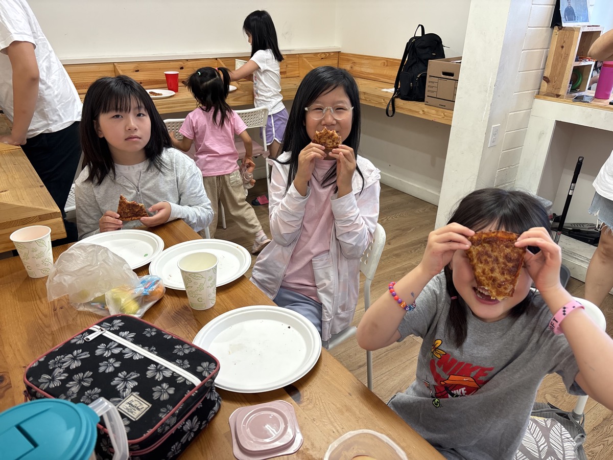 Three kids at the table with pizza slices, playfully covering their eyes