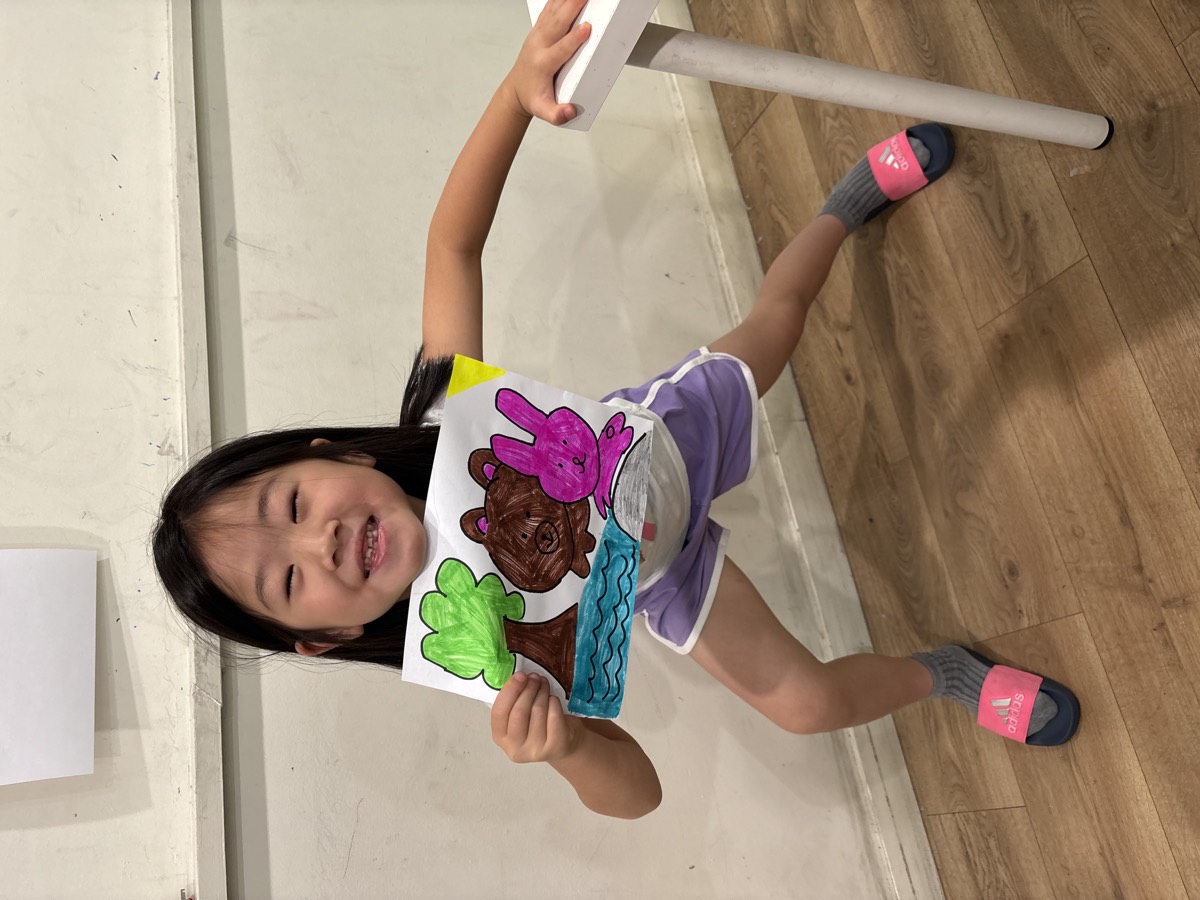 A girl smiling and holding up her finished illustration of a bear and bunny by the water
