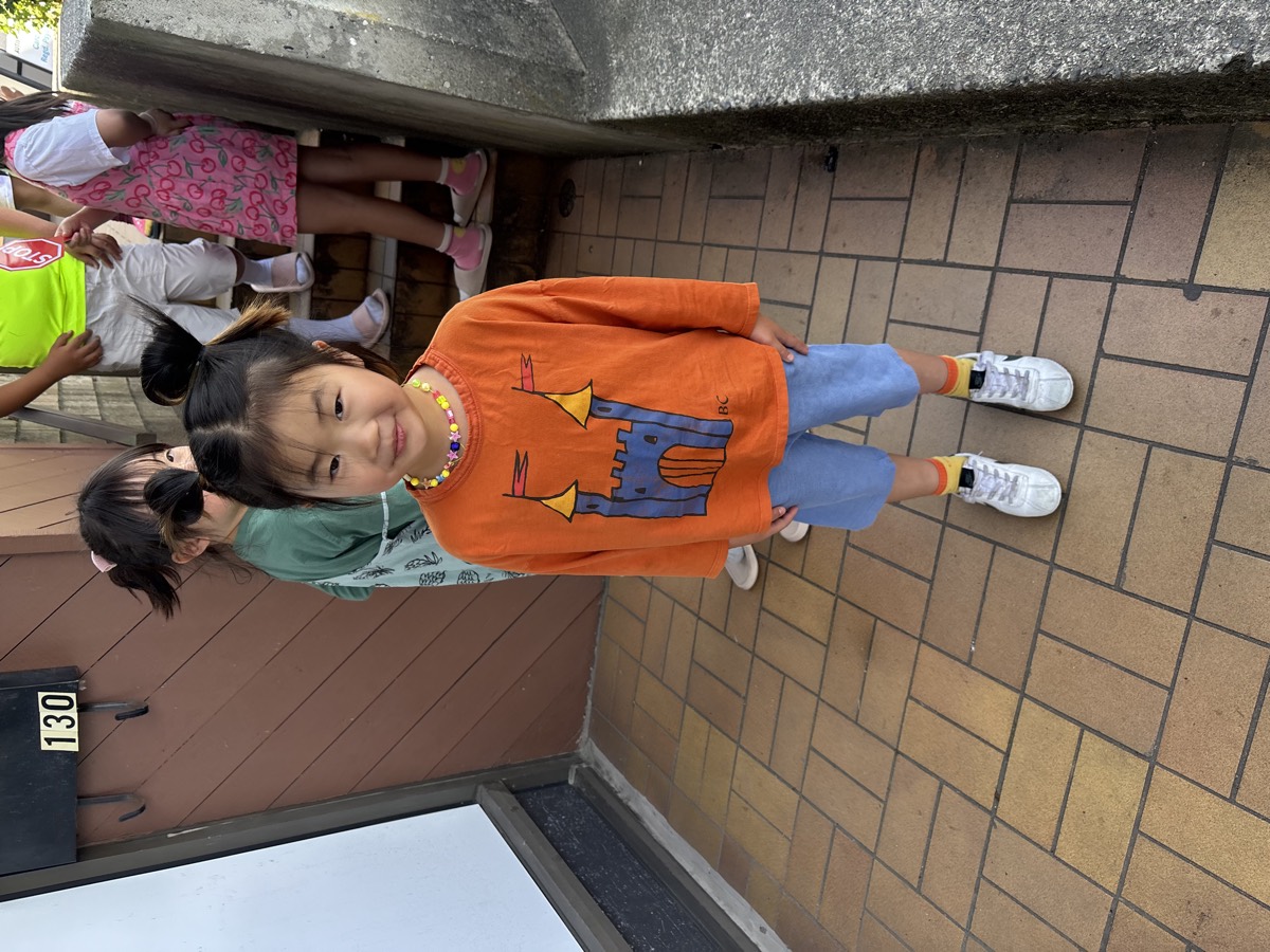 A little girl in an orange castle t-shirt smiling by the school stairs, ready to leave