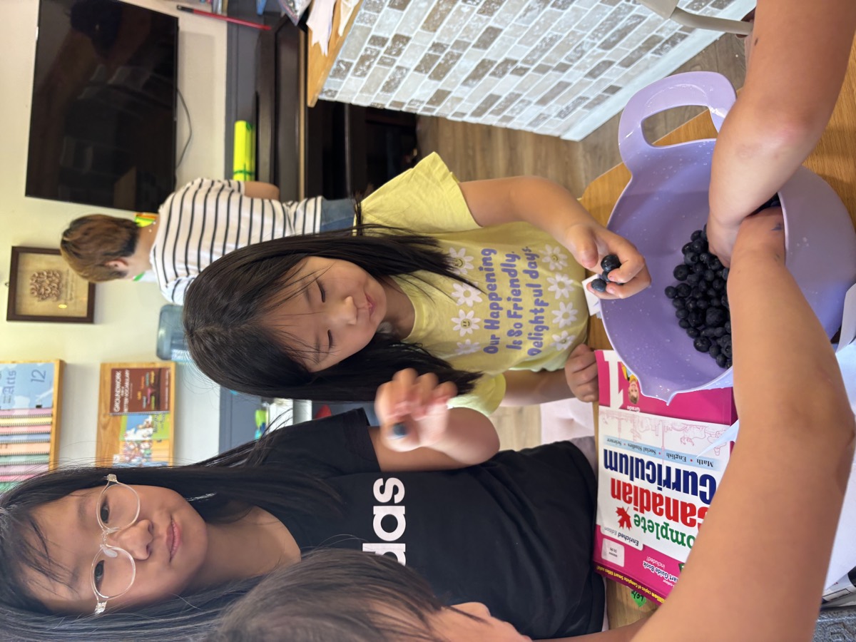 Girls picking blueberries from a purple bowl beside a Canadian Curriculum workbook