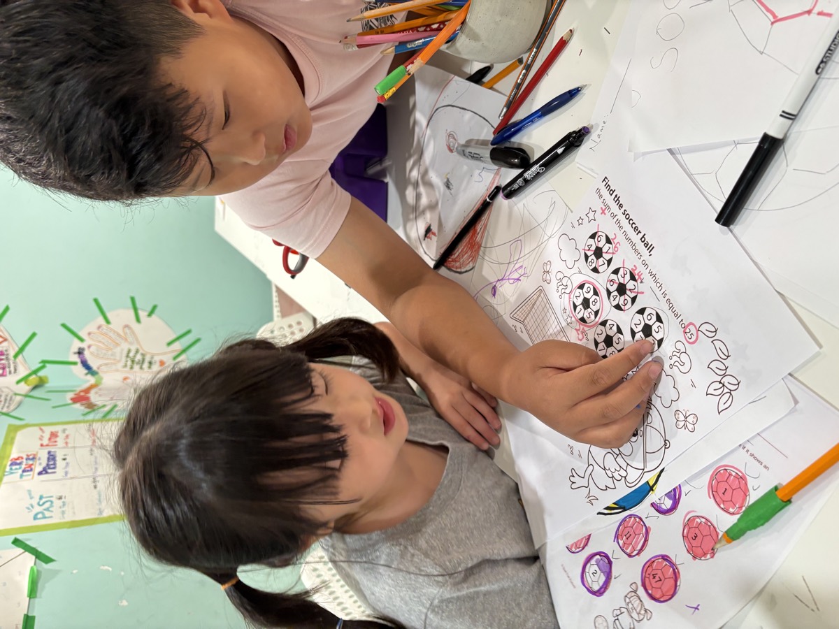 Minwoo leaning over to help a young student with her Find the Soccer Ball worksheet