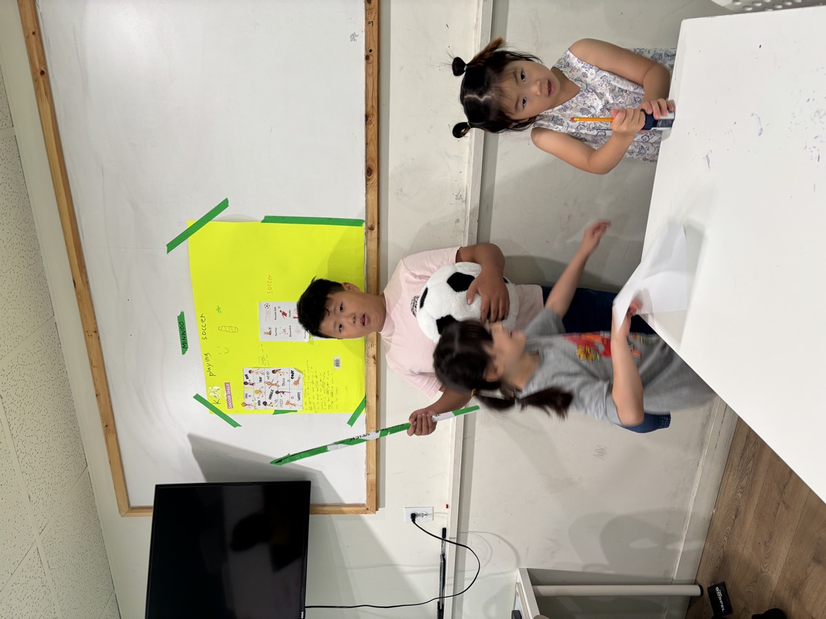 Minwoo standing by his soccer poster with a pointer and ball while two students at the table listen