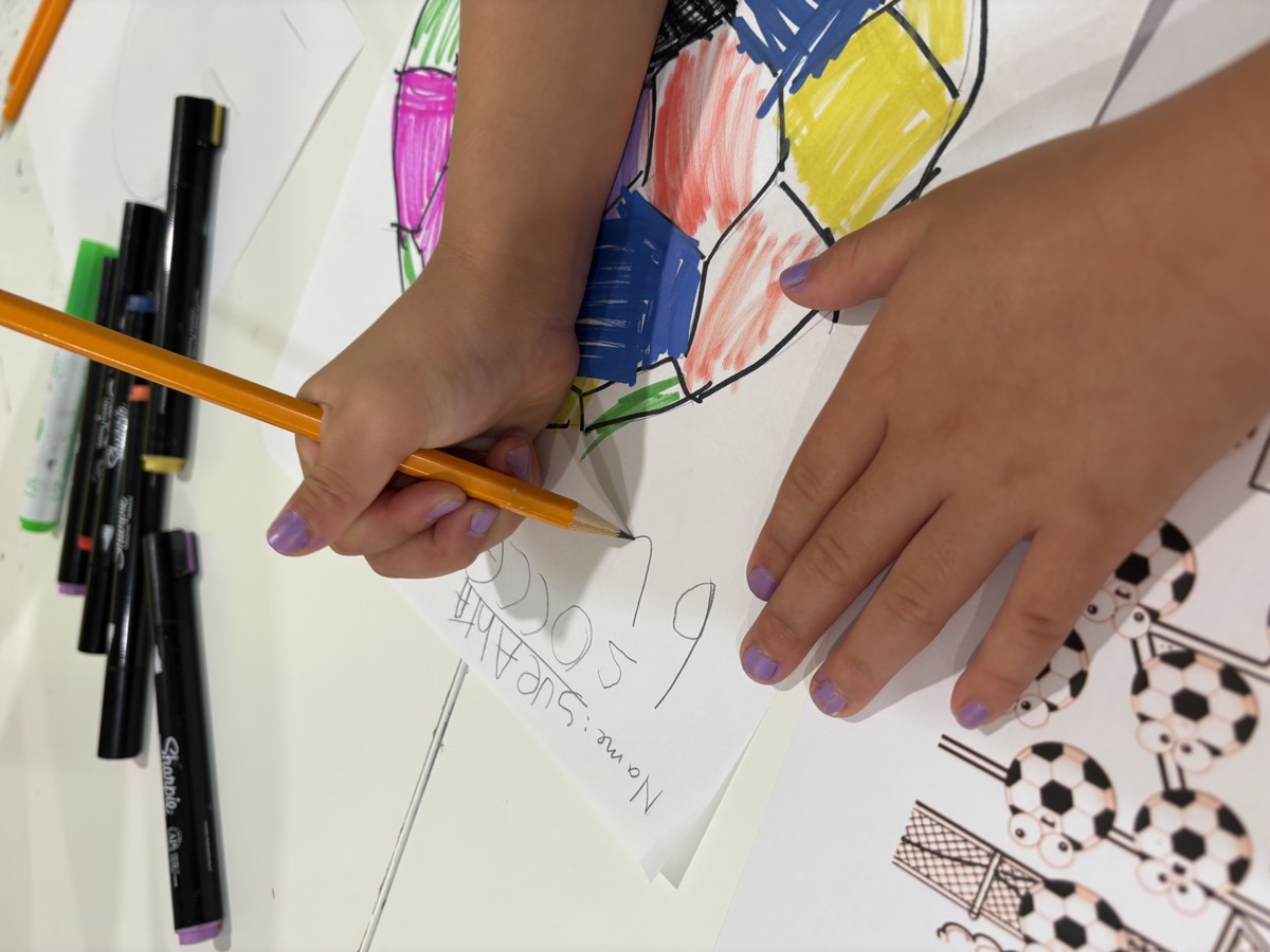 A student writing SOCCER BALL at the top of her worksheet while colouring a soccer ball