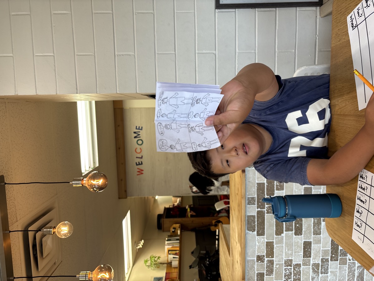An older student holding up his hand-drawn comic-style booklet with speech bubbles under a WELCOME sign