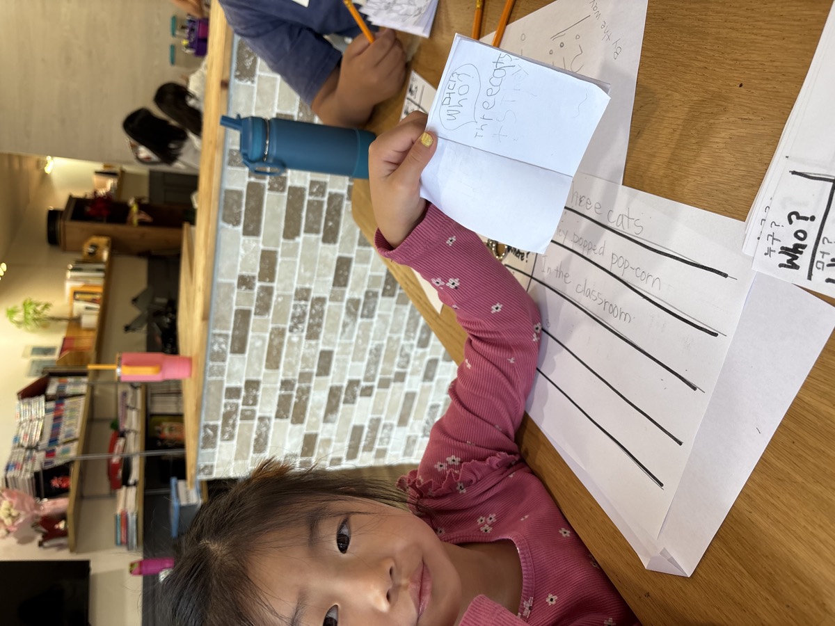 A student in a pink top holding up her mini booklet beside her sentence-strip worksheet