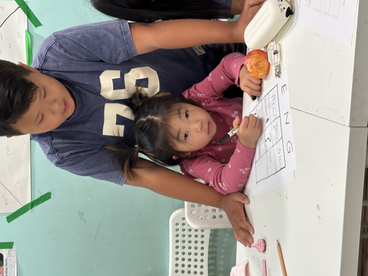An older boy leaning over to help a younger student write letters in the ENG grid on her worksheet