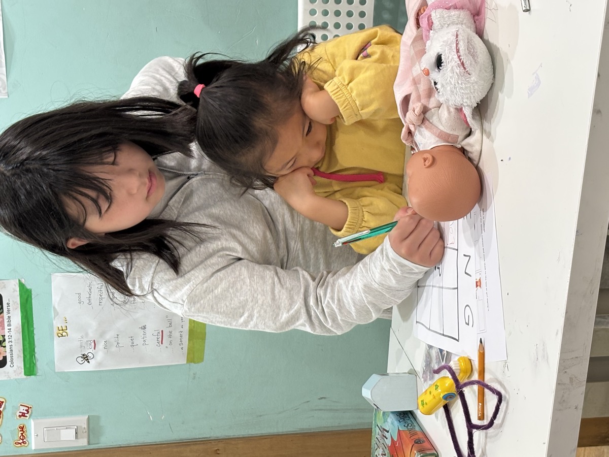 An older girl gently guiding a tired younger child's hand as she writes