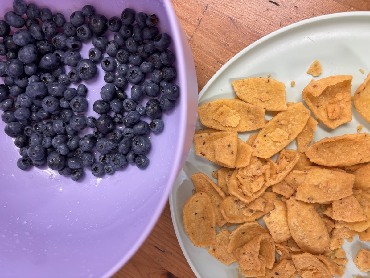 A purple bowl of fresh blueberries beside a plate of crispy corn snacks