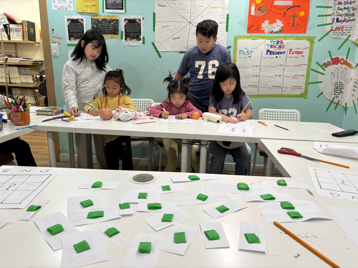 The whole class working at the table with green clay markers on their ENG grid sheets, verb tense charts on the wall behind