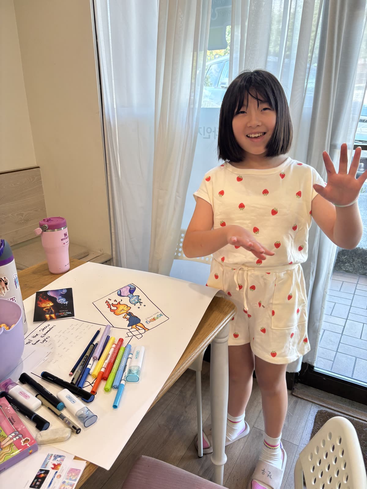 A young student standing proudly beside her drawings and markers