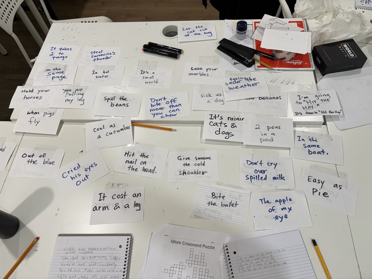 Dozens of handwritten idiom cards spread out across the table, including spill the beans, bite the bullet and easy as pie