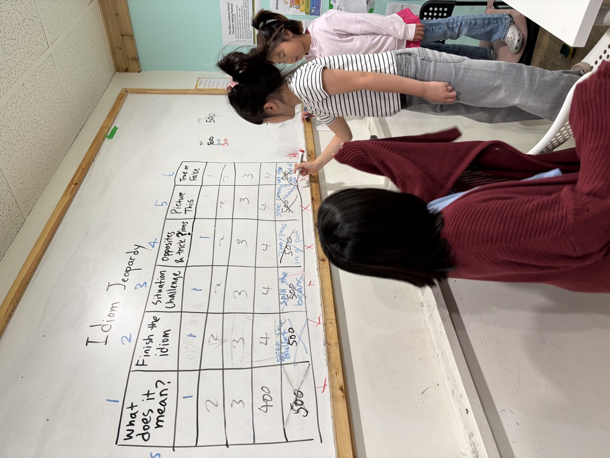 Three students at the whiteboard playing Idiom Jeopardy, one pointing at a category cell in the big scoring grid