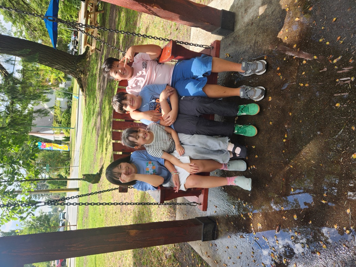 Four students sitting together on a wooden bench swing under a shelter at the park, smiling
