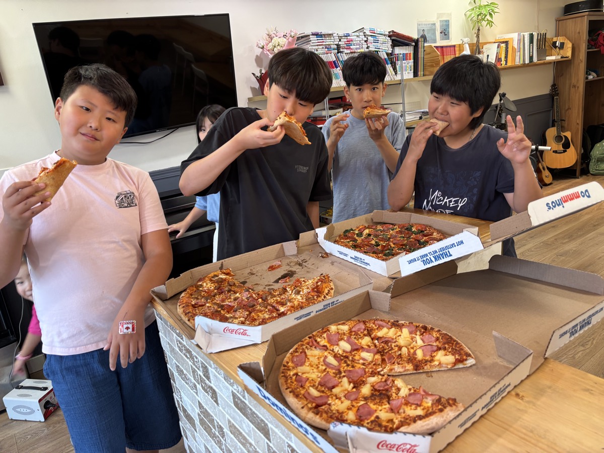 Four boys smiling and eating pizza together around a table stacked with pizza boxes, one making a peace sign