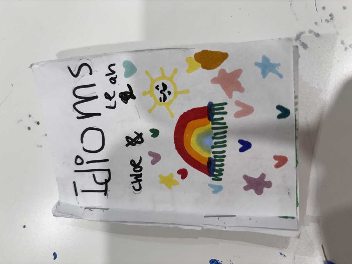 A handmade booklet cover reading Idioms by Chloe and Leah, decorated with a rainbow, sun, stars and hearts