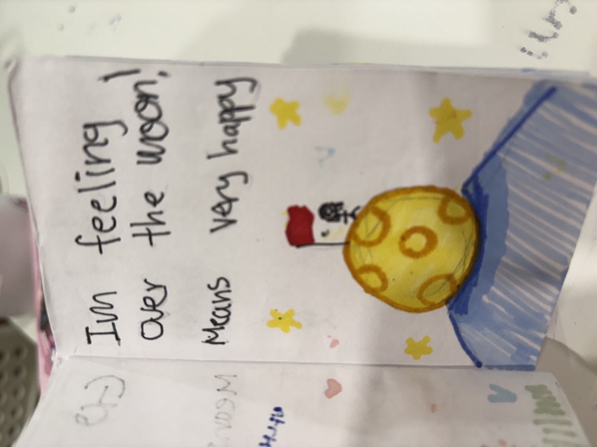 A booklet page reading I'm feeling over the moon means very happy, with a drawing of a figure standing on the moon