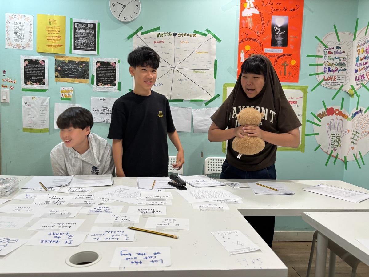Three students standing at the front of the classroom during Idiom Jeopardy, one holding a teddy bear, with idiom cards spread across the table
