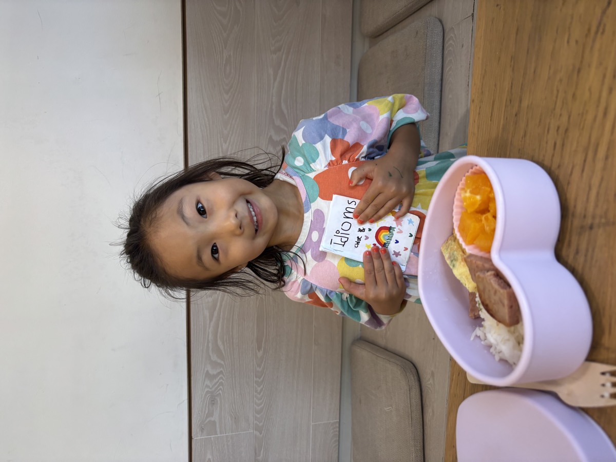 A young student smiling and holding up her handmade idiom booklet at the lunch table beside her lunch tray