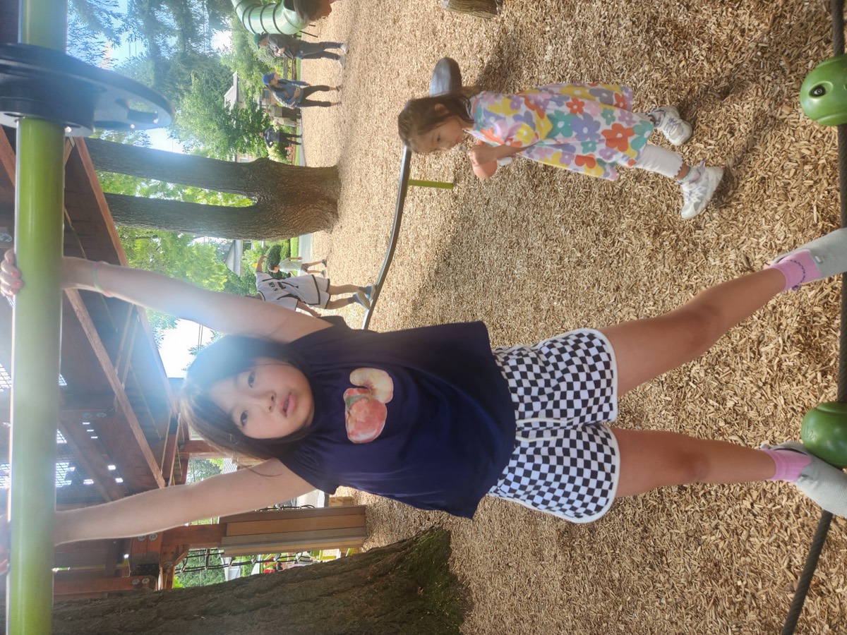A student hanging from the overhead bar of the wooden play structure, smiling at the camera