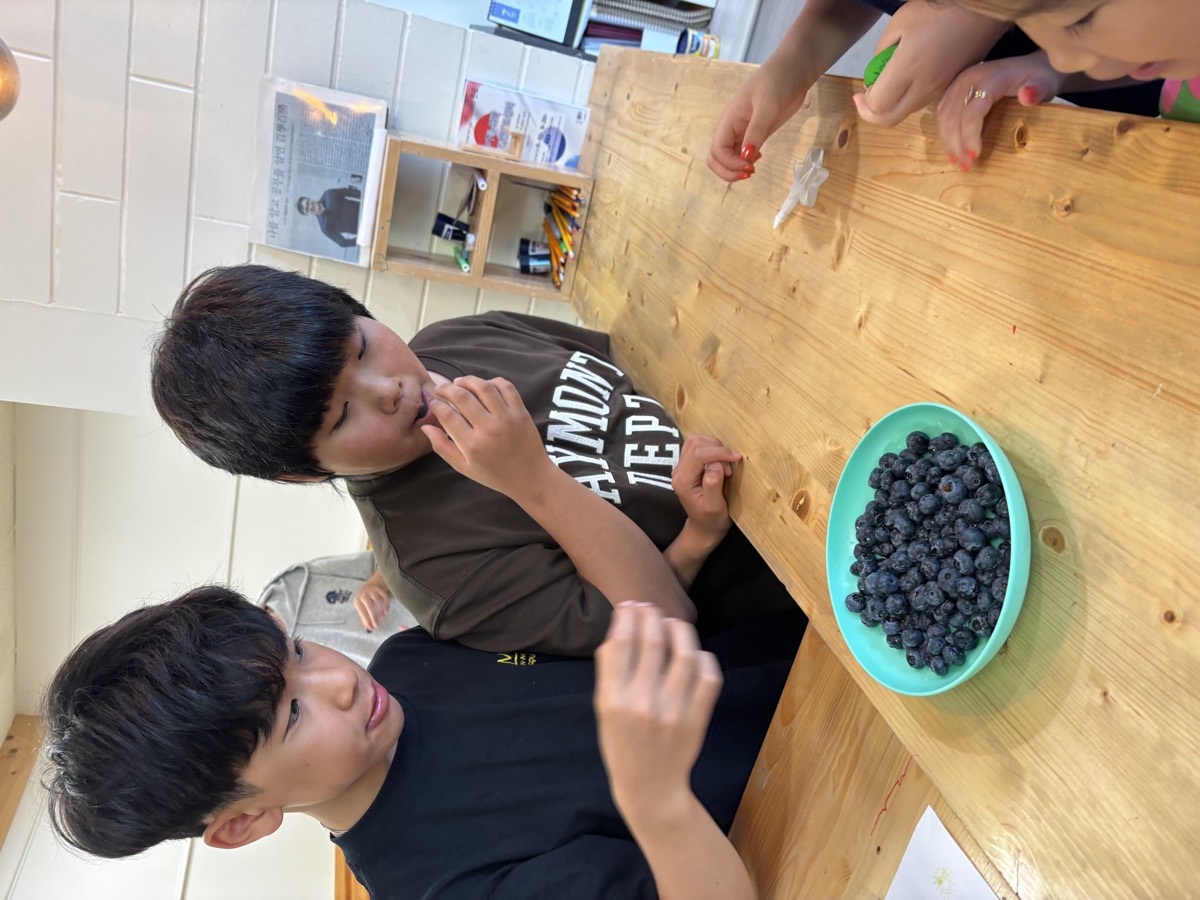 Two students eating fresh blueberries from a green bowl on the classroom table
