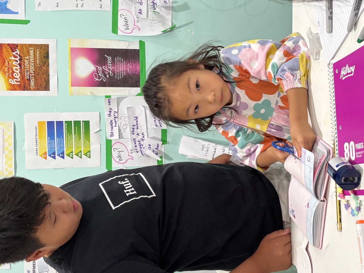 An older student standing beside a much younger student, helping her with the open notebook on her desk