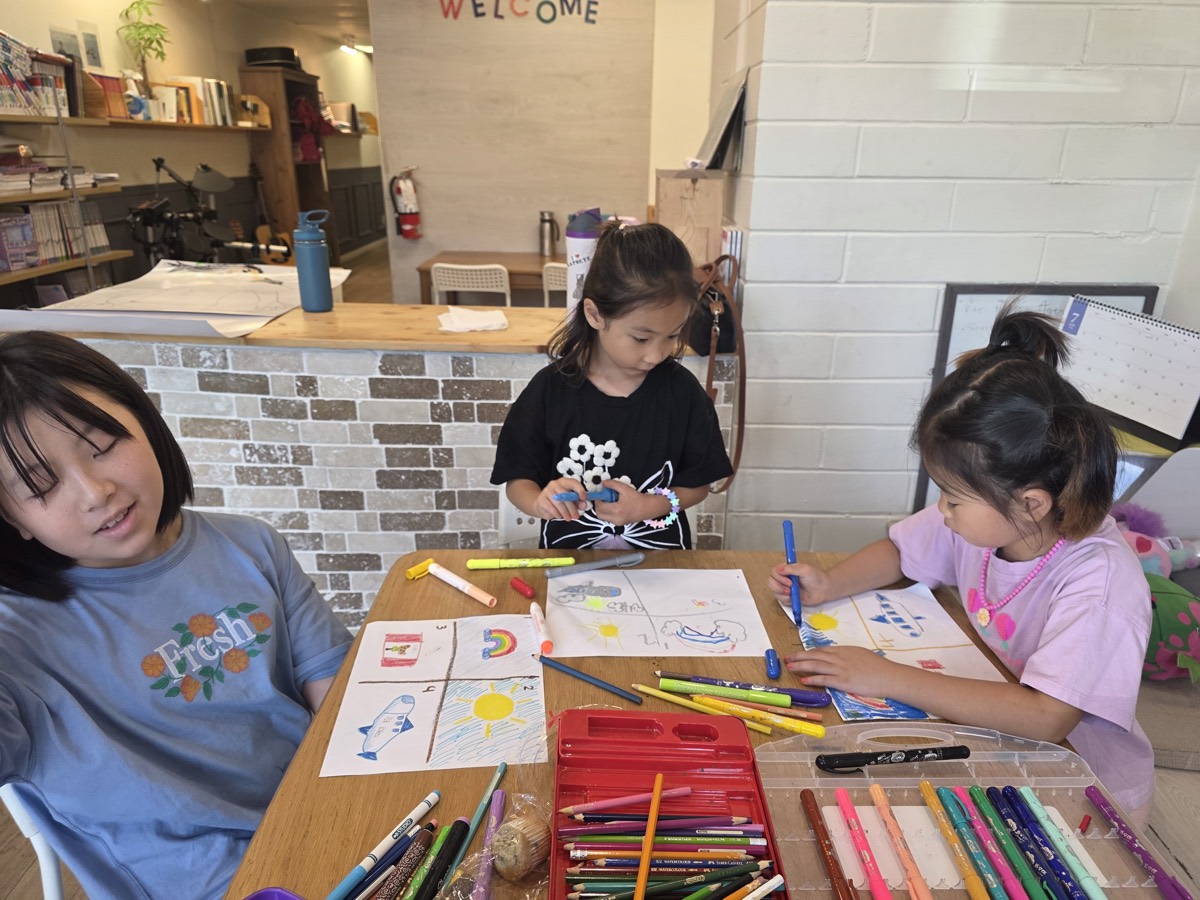 Three students drawing at the table with a long row of coloured markers laid out in front of them