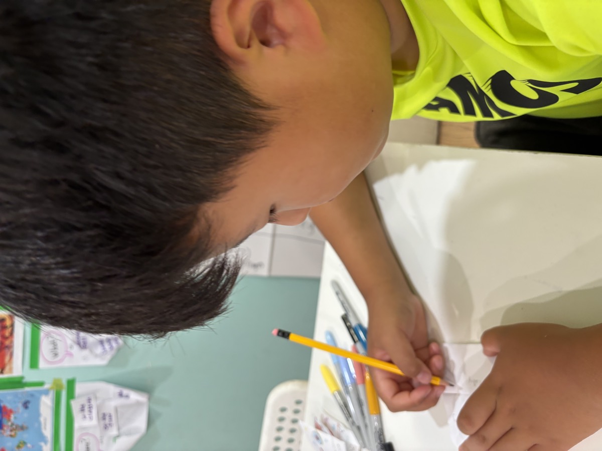 A student in a bright yellow shirt leaning over the table, writing carefully in his small flip-book