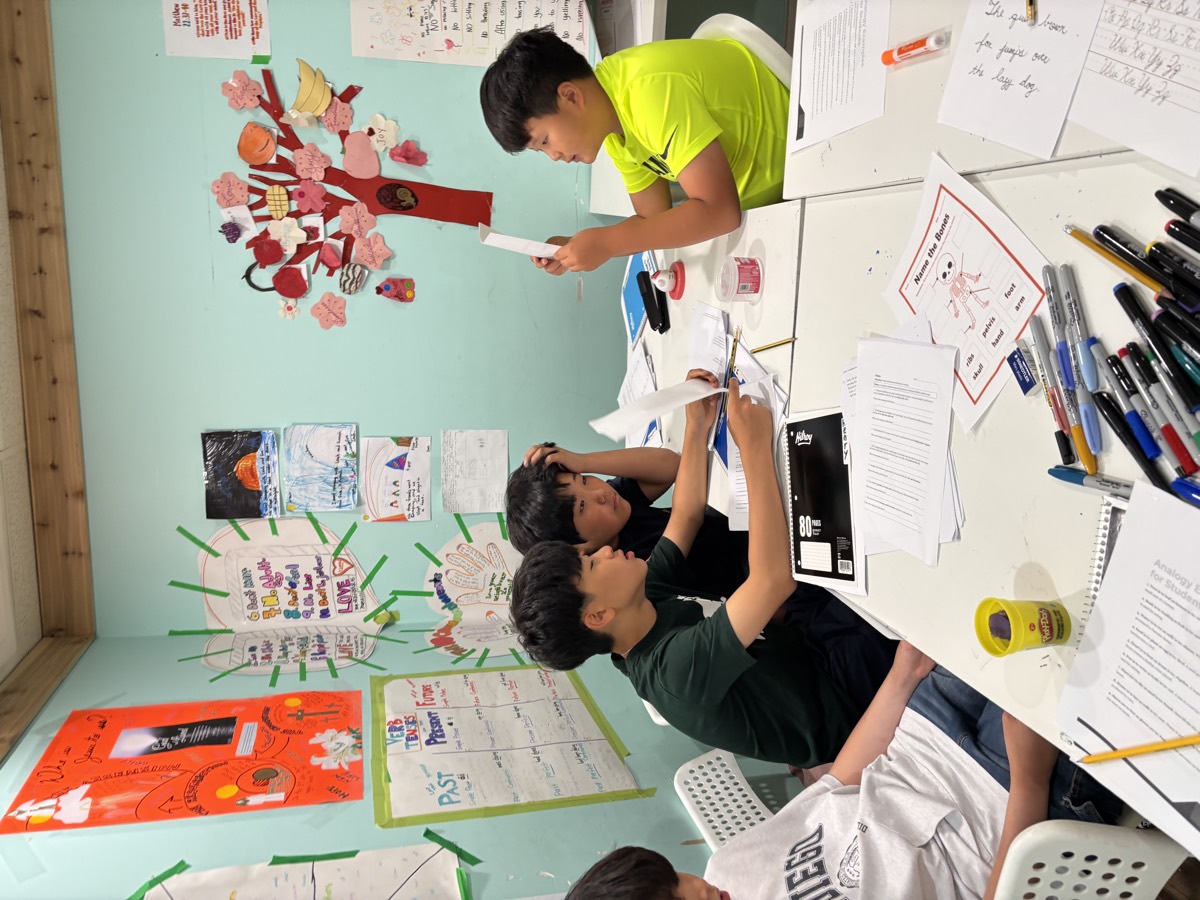 Middle school students seated around the table, one reading aloud from a sheet of paper while the others listen