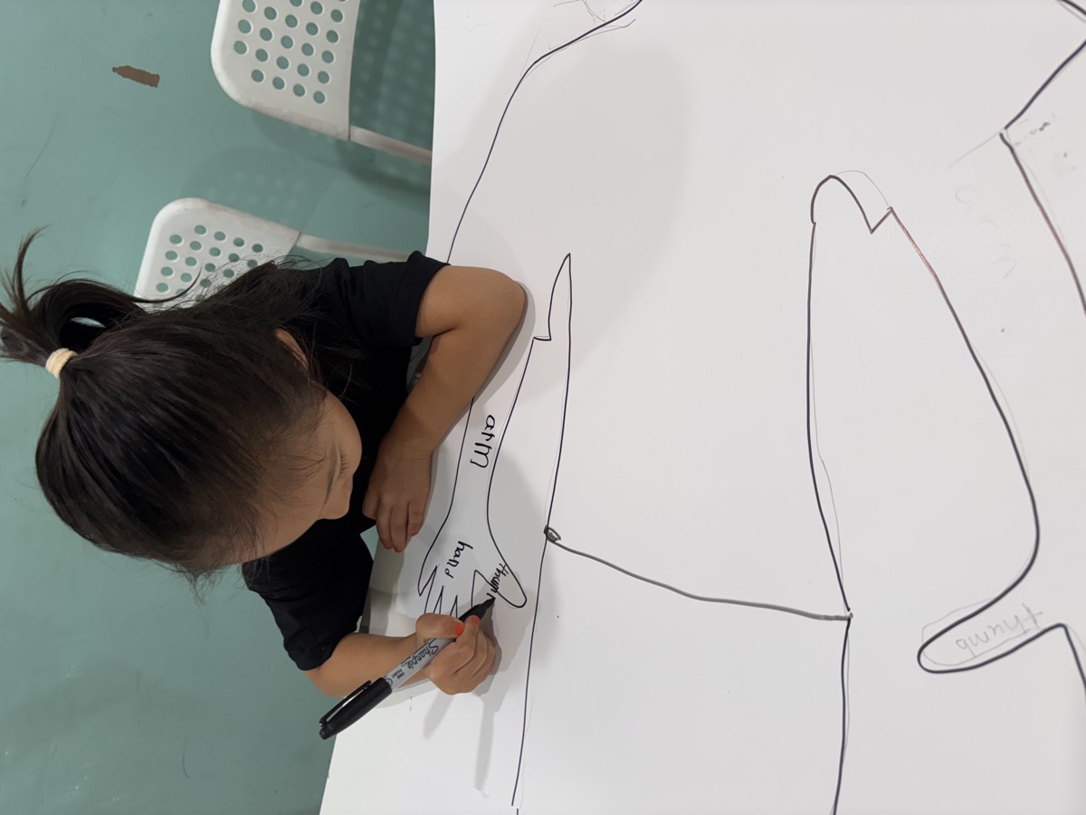 A student leaning over the large traced body outline on the table, writing on it with a black marker